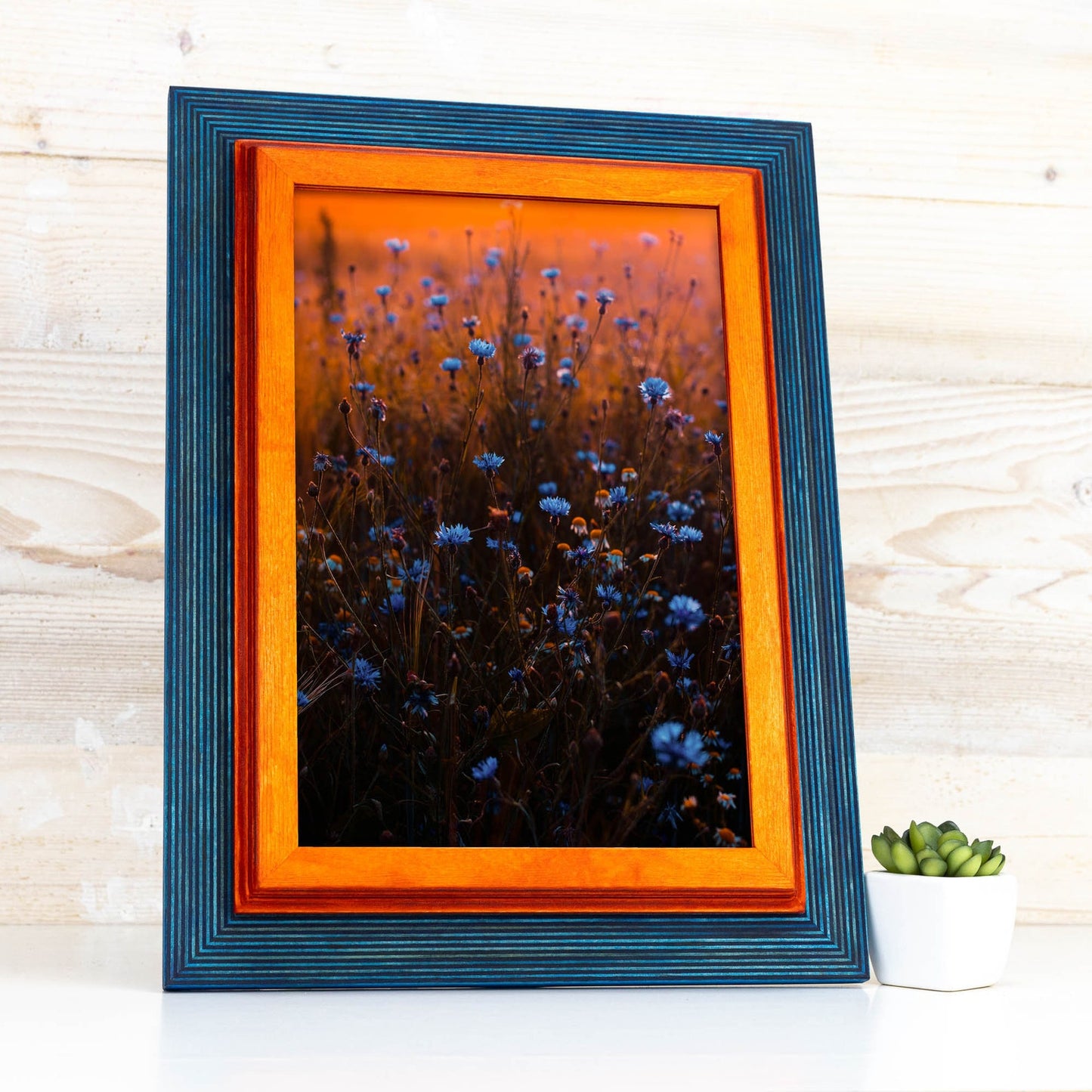 Framed artwork of blue flowers with an orange border on a wooden surface. Add rustic elegance with this grey + white birch frame. Handmade and built to last, ideal for artwork, photo décor, or Scandi-inspired homes.