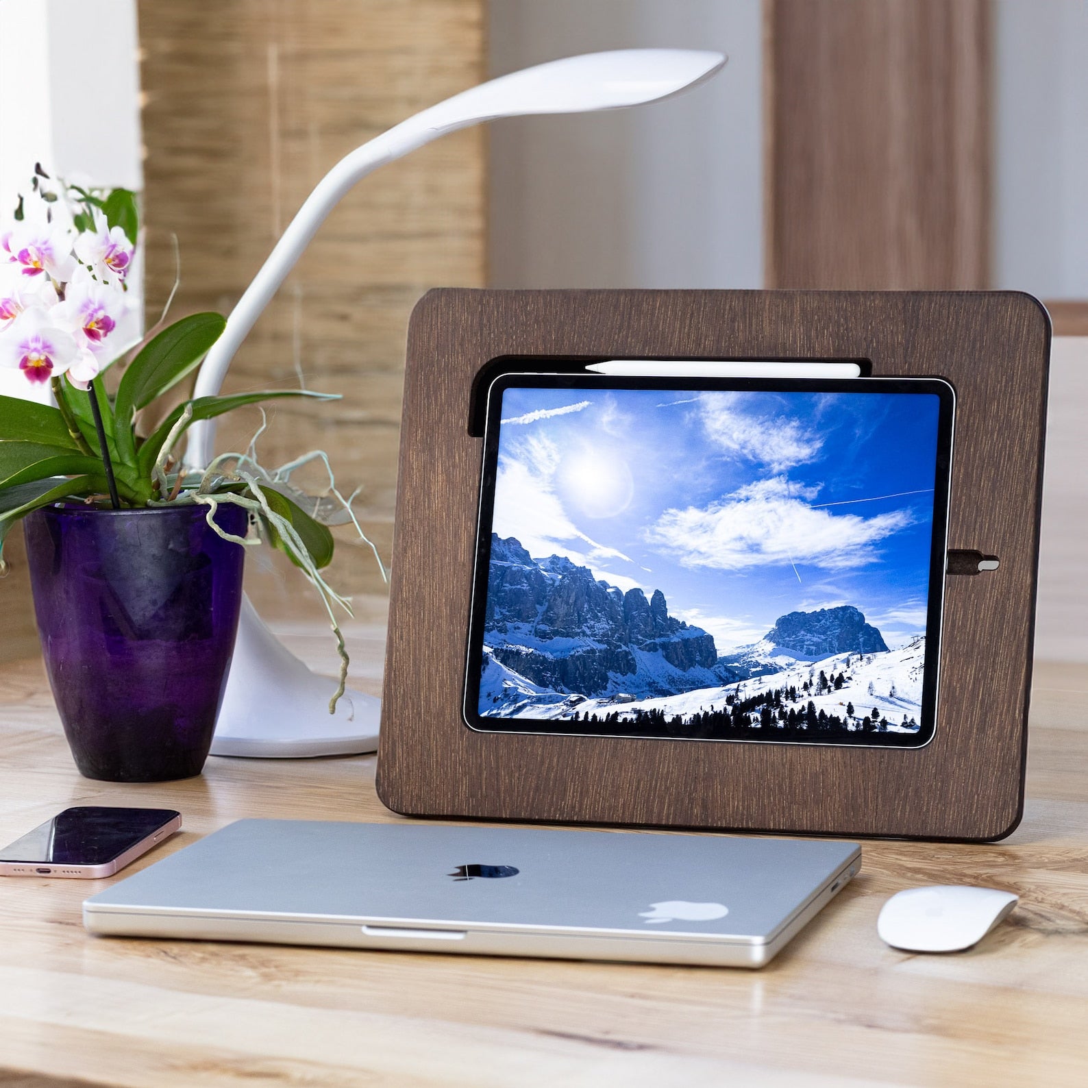 A dark brown wooden stand holding an iPad placed on a table next to Apple products and a flower