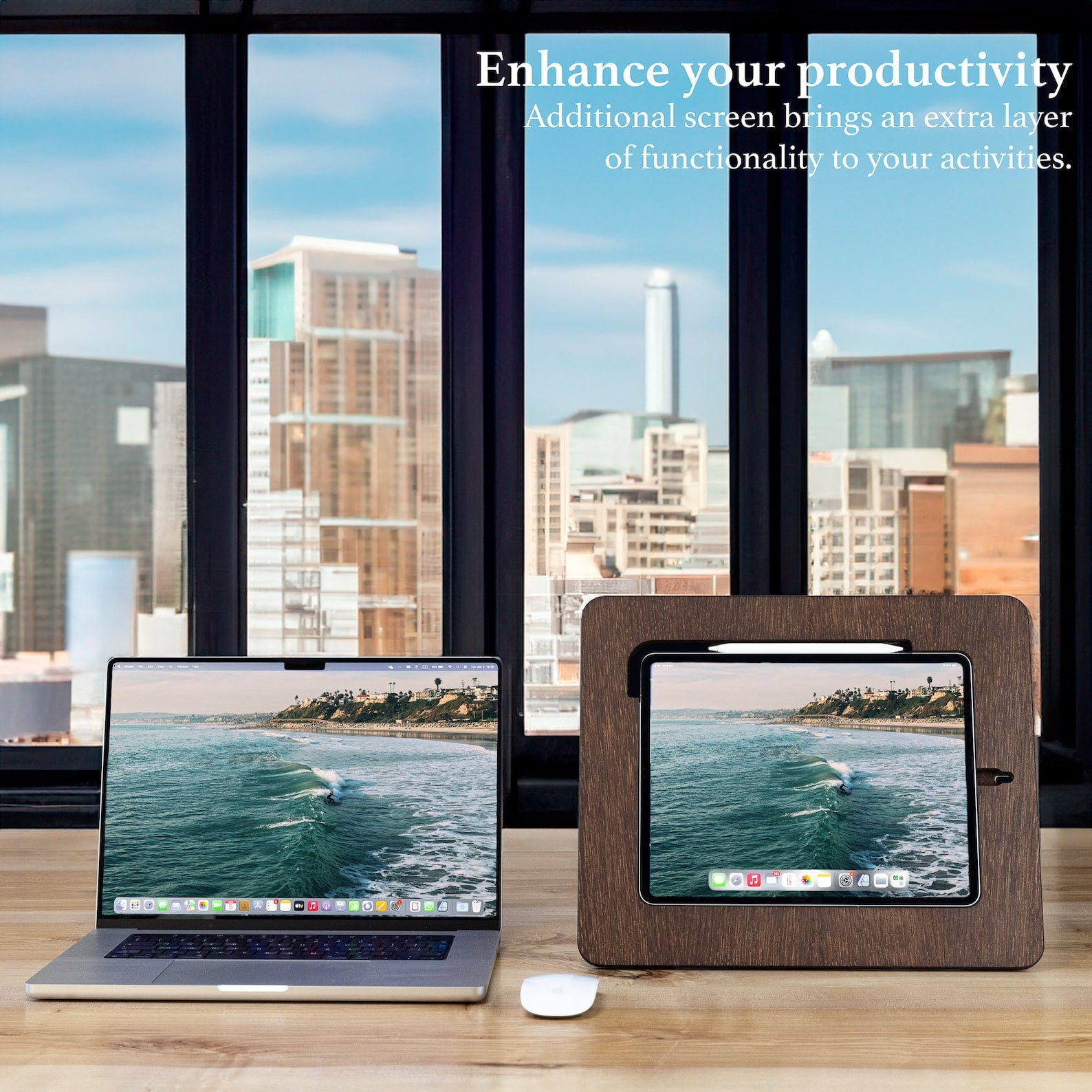 A dark wooden board holding an iPad and an Apple pen placed next to a laptop on the table overlooking a view of a city.