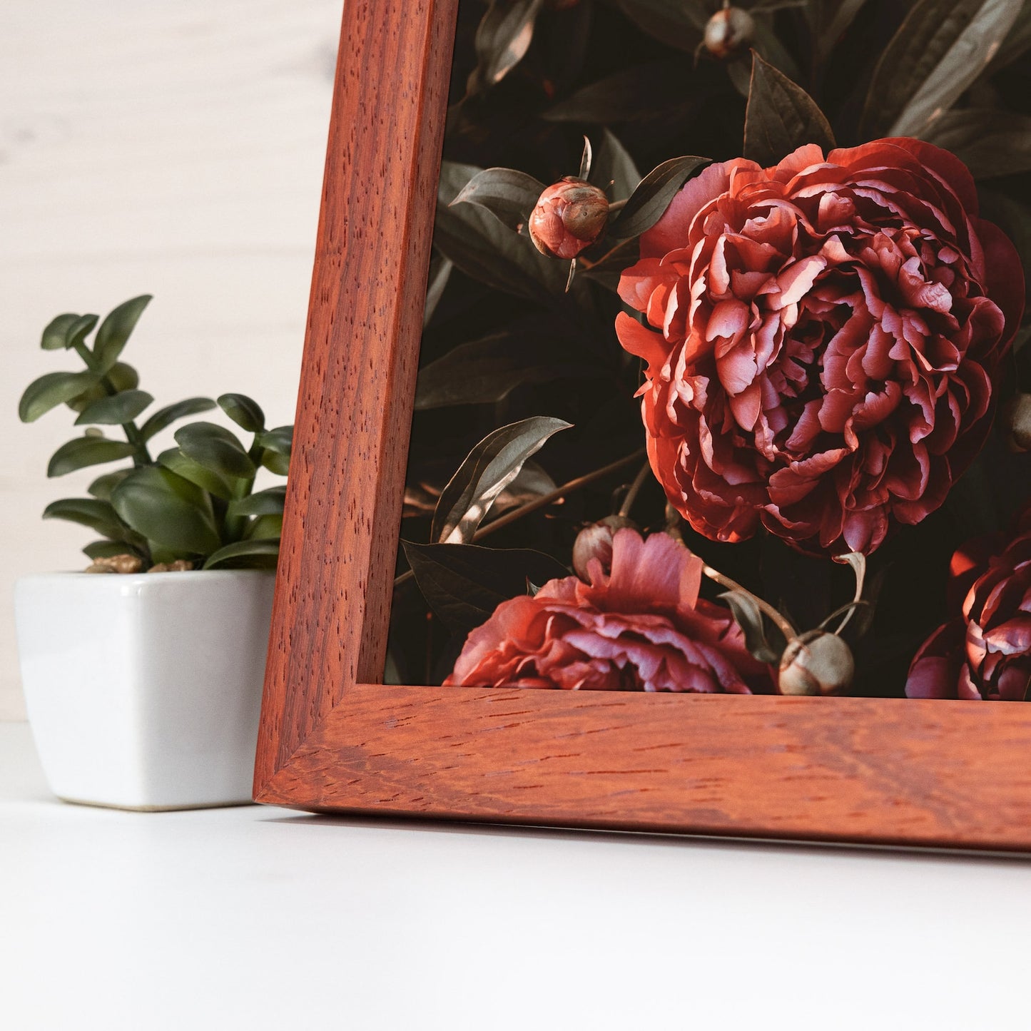 Wooden frame with a floral arrangement on a white surface with a plant in the background.Handmade alder hardwood frame with rich brown tones and red accents. Finished in natural oil, it adds warmth and charm to your artwork or photos.