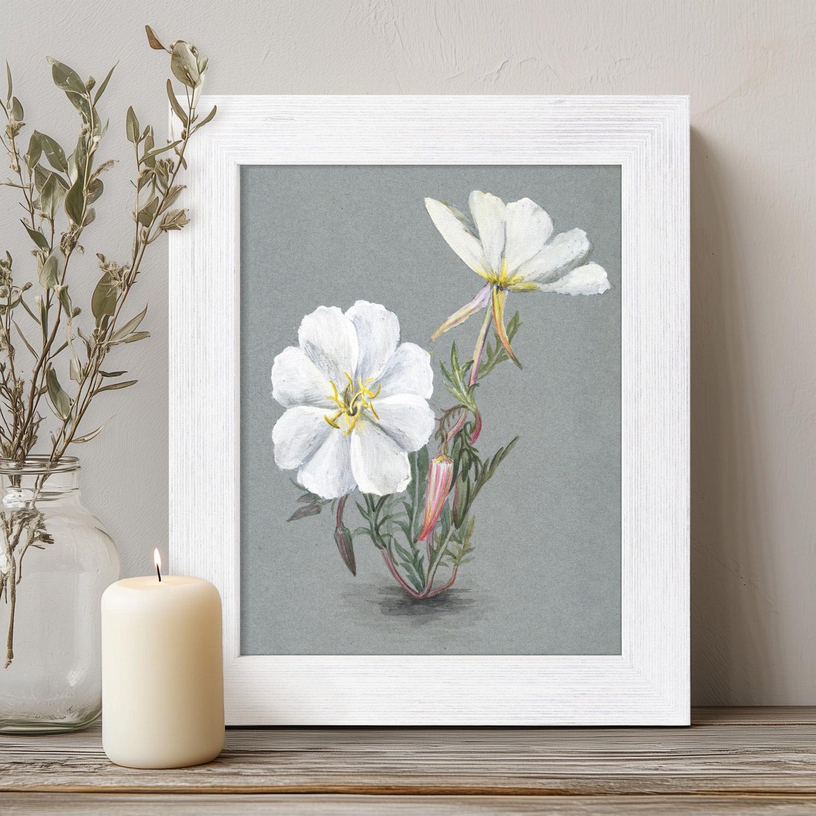 Framed botanical print of white flowers on a gray background, placed on a wooden surface with a candle and vase.