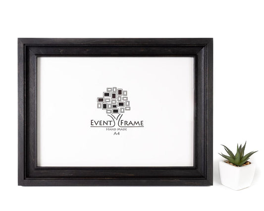A black vintage design photo frame made of solid birch hardwood, displayed with a white mat and a small potted plant to the side.