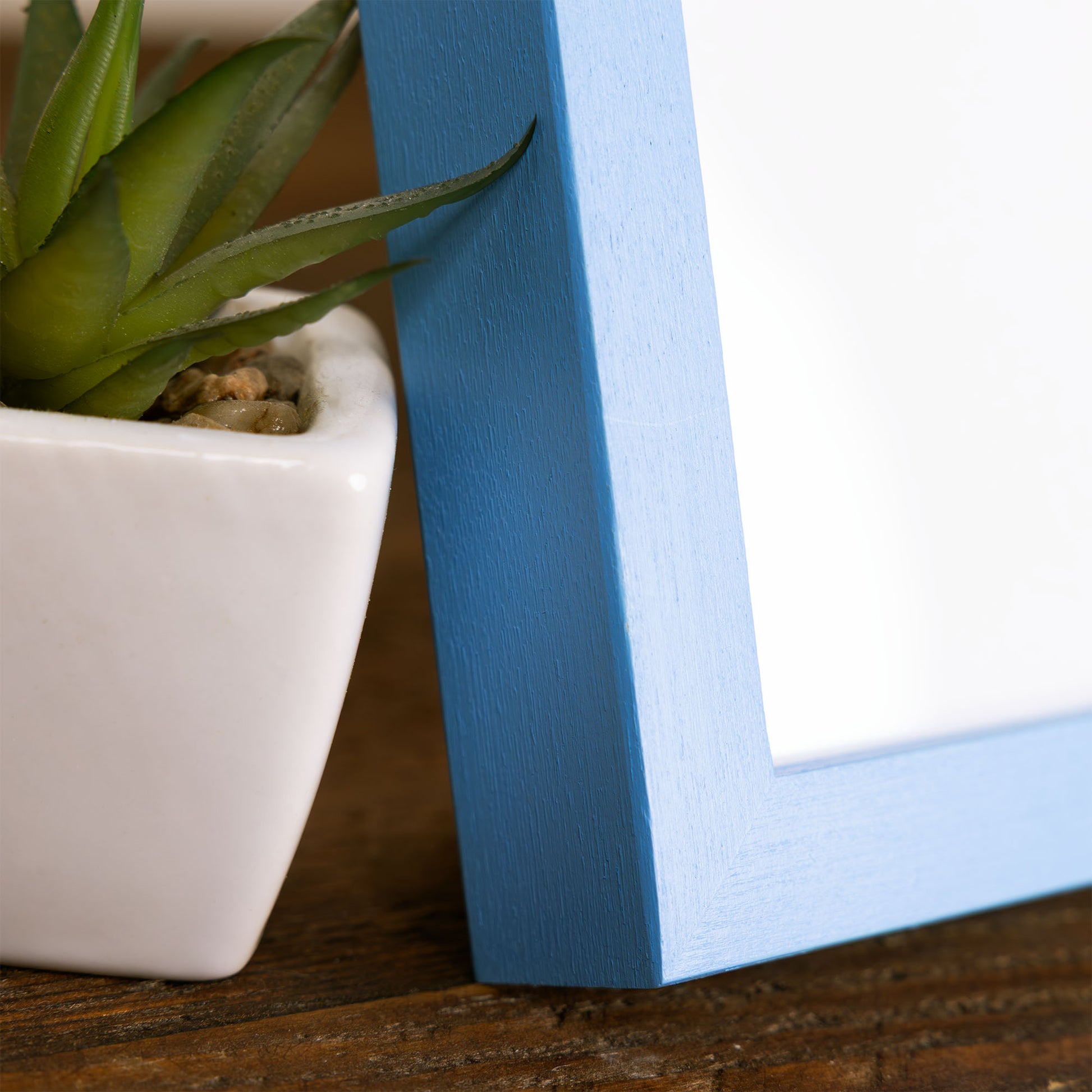 Blue framed picture frame leaning against a white pot with a green plant on a wooden surface