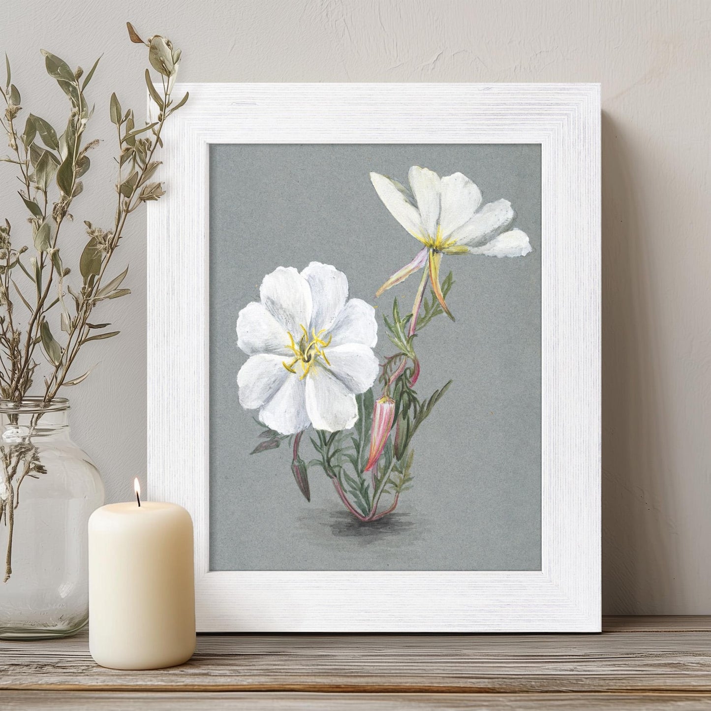Framed botanical print of white flowers on a gray background, placed on a wooden surface with a candle and vase.