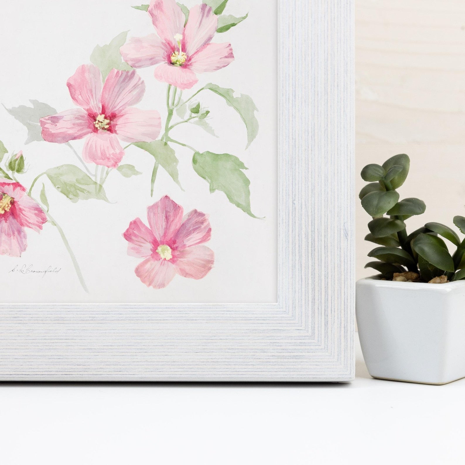 Framed floral artwork with pink flowers on a white surface next to a potted plant.